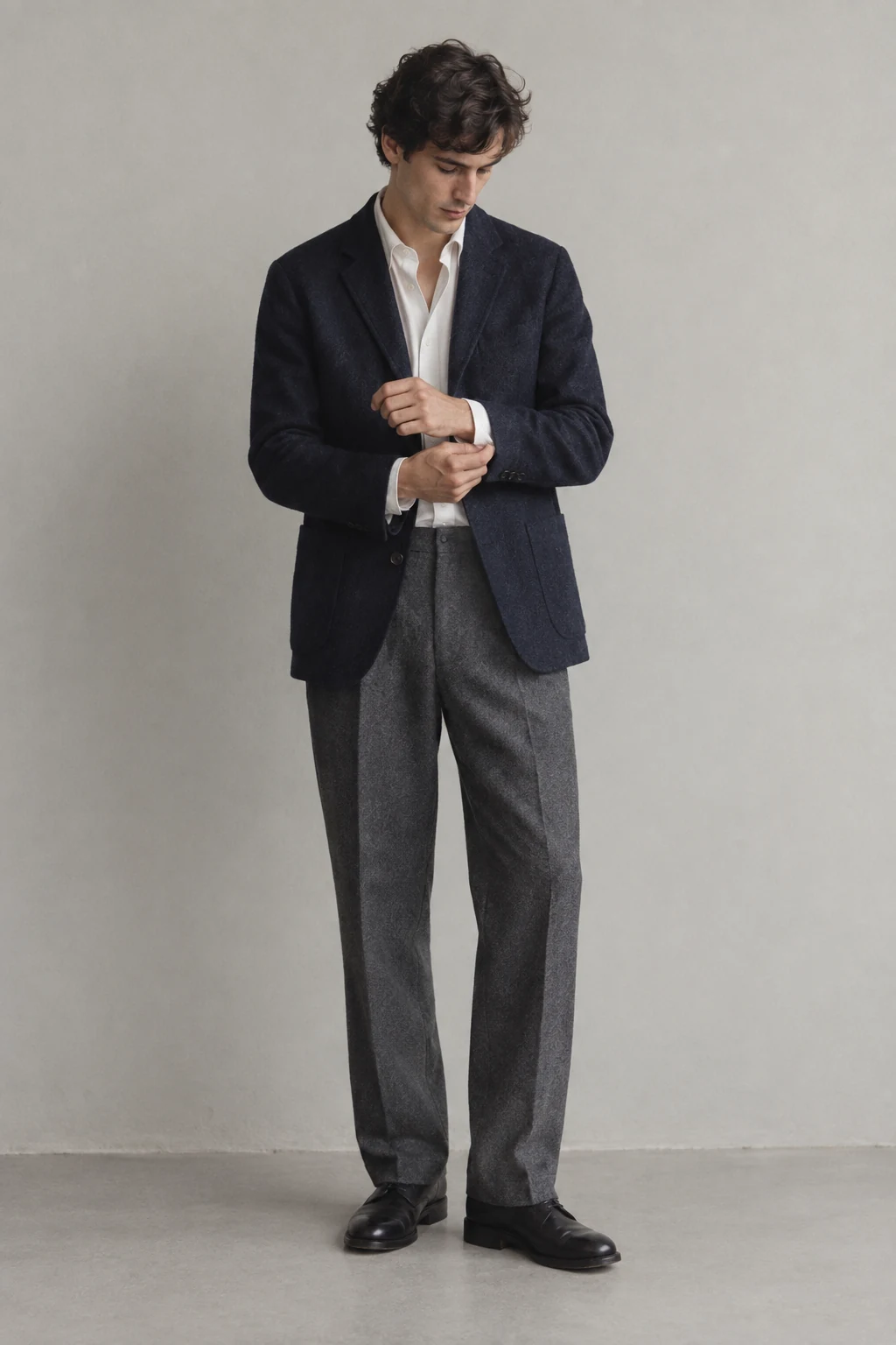 A young man in a navy wool jacket and gray trousers looks down while adjusting his white shirt cuff.