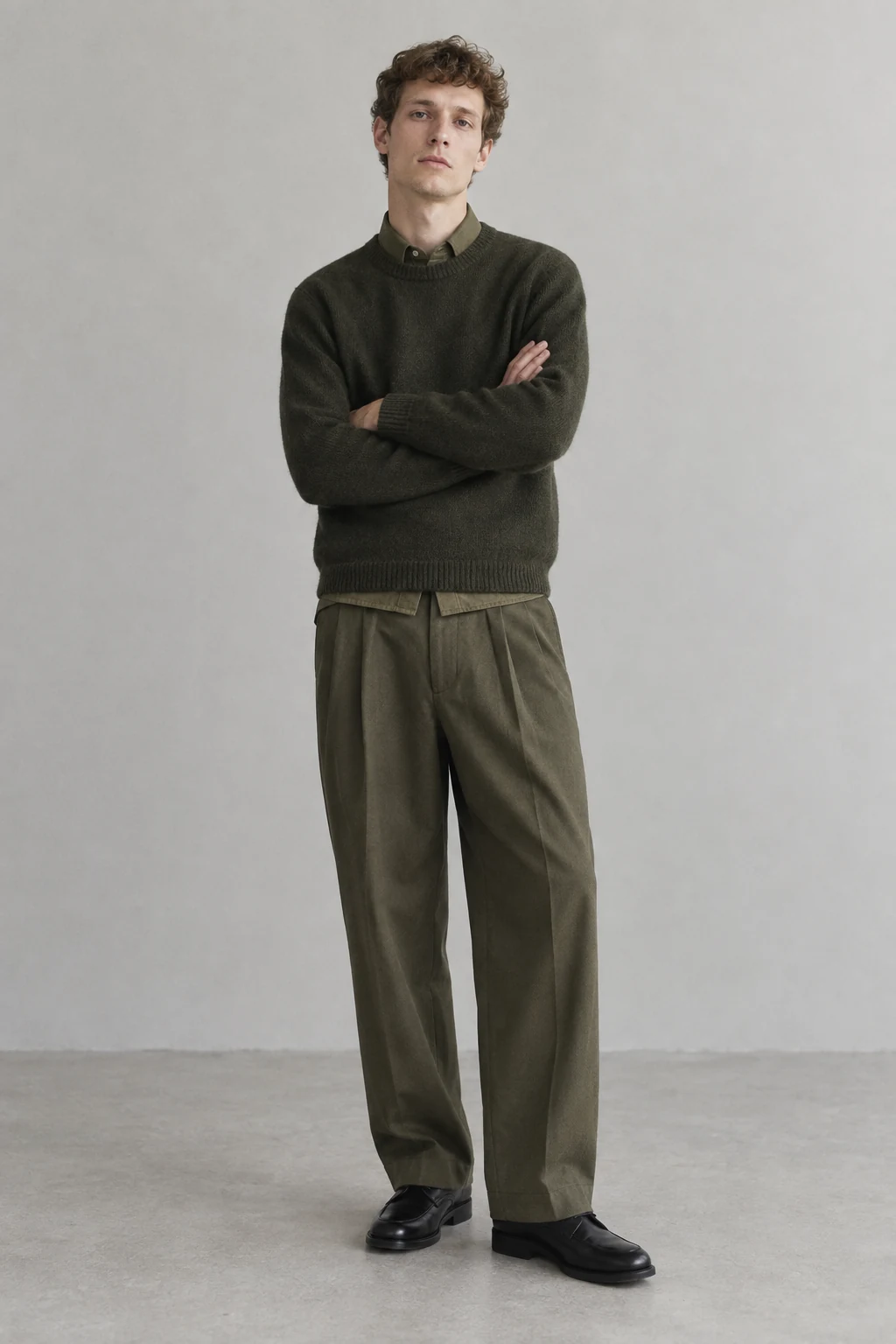 A young curly haired man stands with folded arms in layered olive knitwear wide pleated trousers and black leather shoes.
