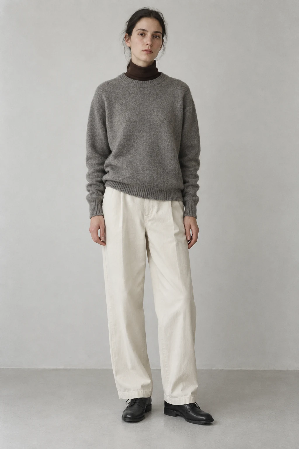 A young woman stands in a gray sweater, brown roll neck, wide cream trousers, and black lace up shoes.