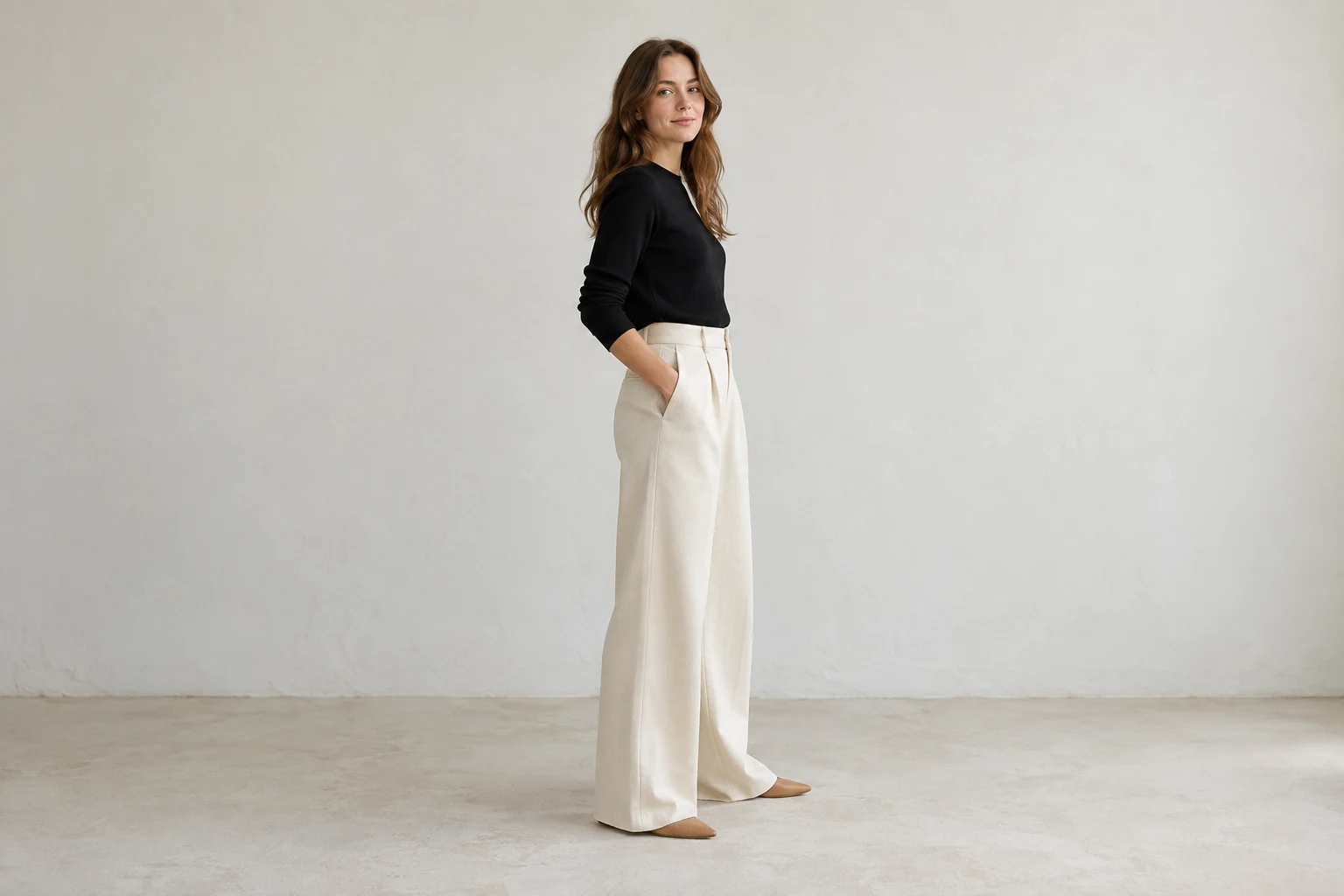 A woman stands in cream wide leg trousers with a black knit top and tan pointed flats against a pale plaster wall.