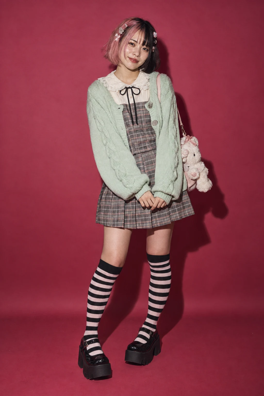 A young Japanese woman poses against raspberry pink in a mint cardigan, checked pinafore, striped socks and platform shoes.